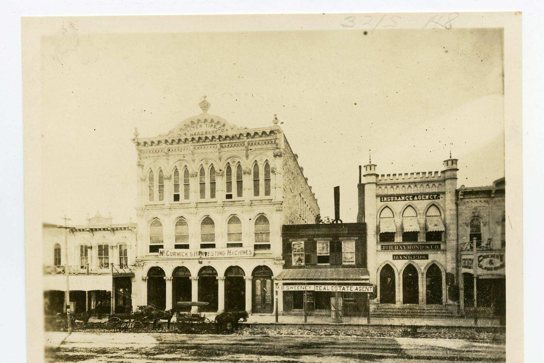 A mounted sepia photograph of a row of commercial buildings on an unpaved street, with signs for harvesting machines, a real estate agent, an insurance agency and bankers, horse drawn wagons at the curb and rails in the roadway.