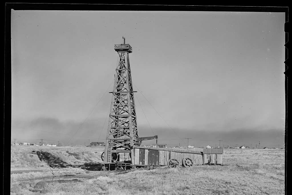 A black and white photograph of a tall wooden oil derrick beside a weathered plank shed on a wheeled frame, set on open grassland with low houses along the horizon.