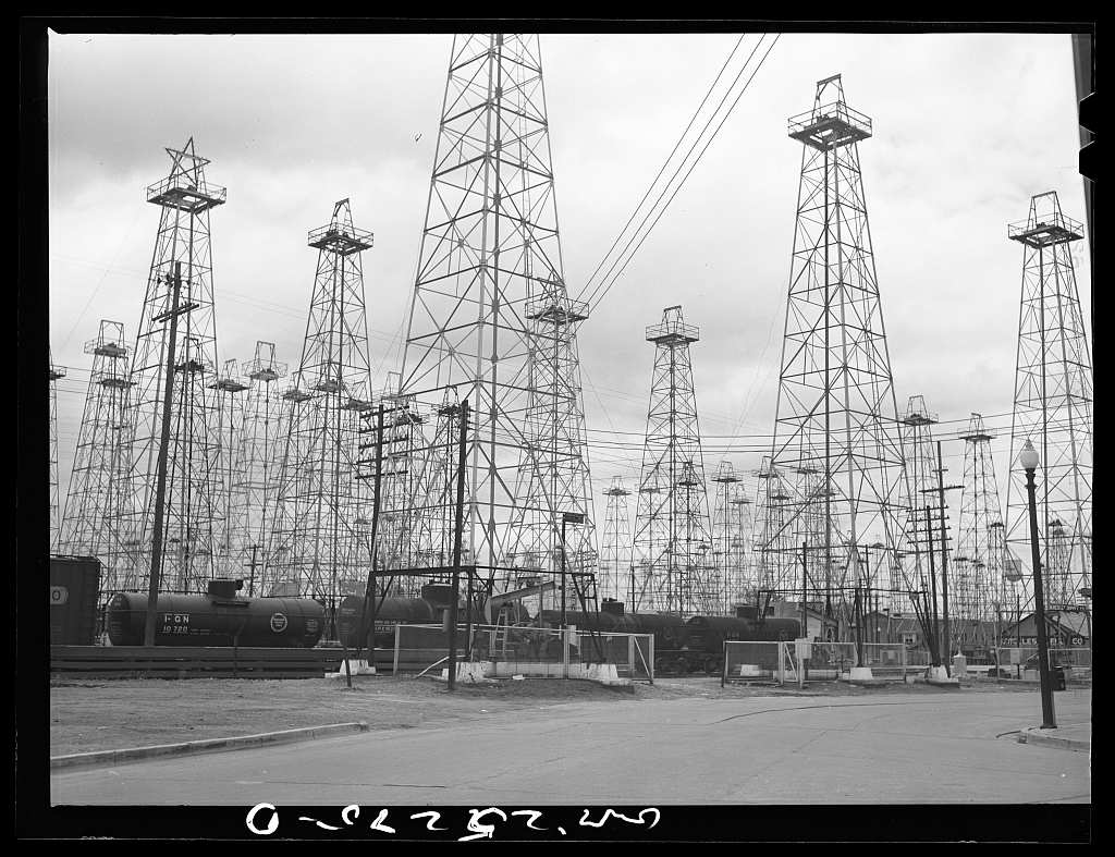 Dozens of steel oil derricks crowd a block in Kilgore behind a row of railroad tank cars and a low fence, with a street lamp and an empty paved road in the foreground.