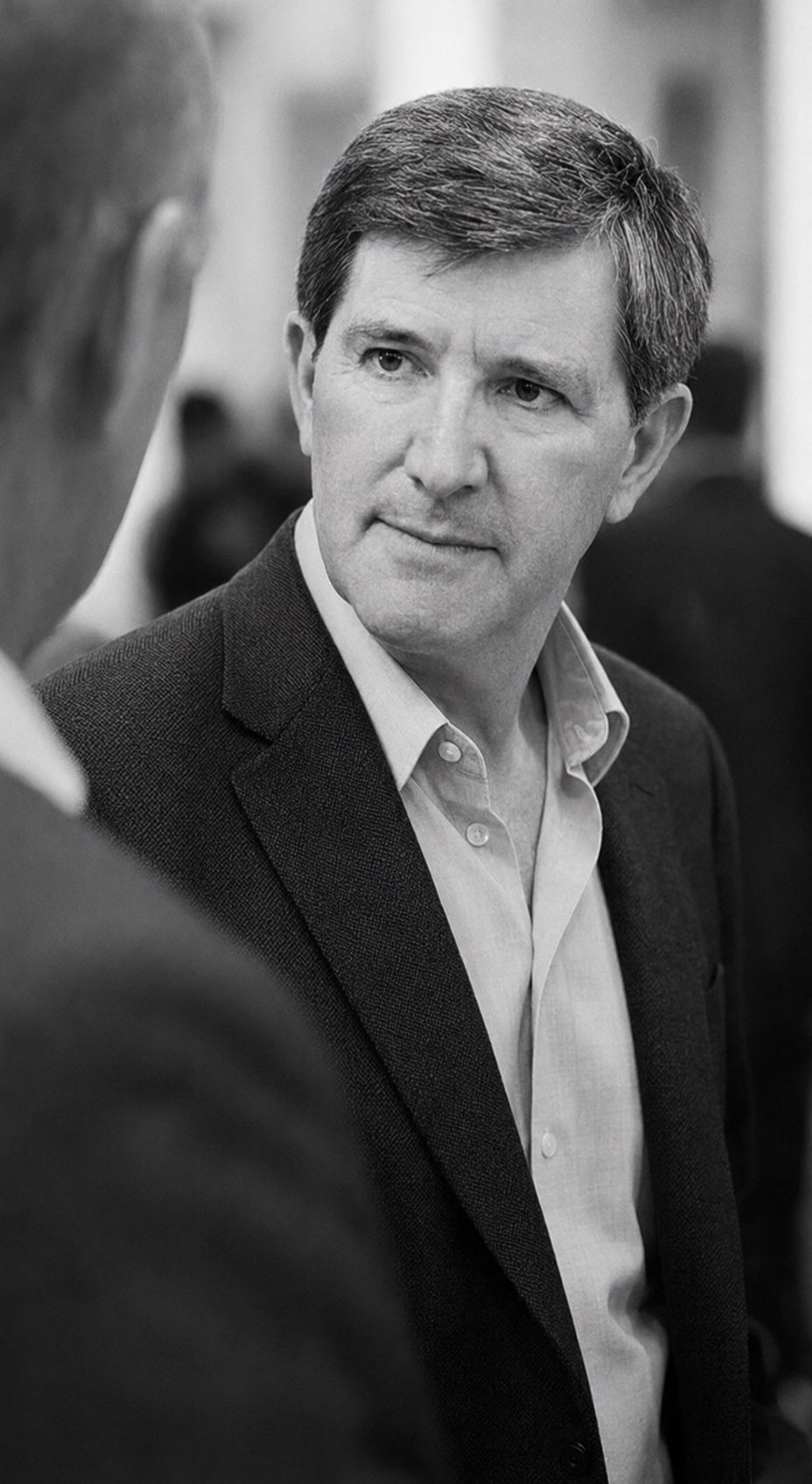 Black and white photograph of him listening at a gathering, another guest seen from behind